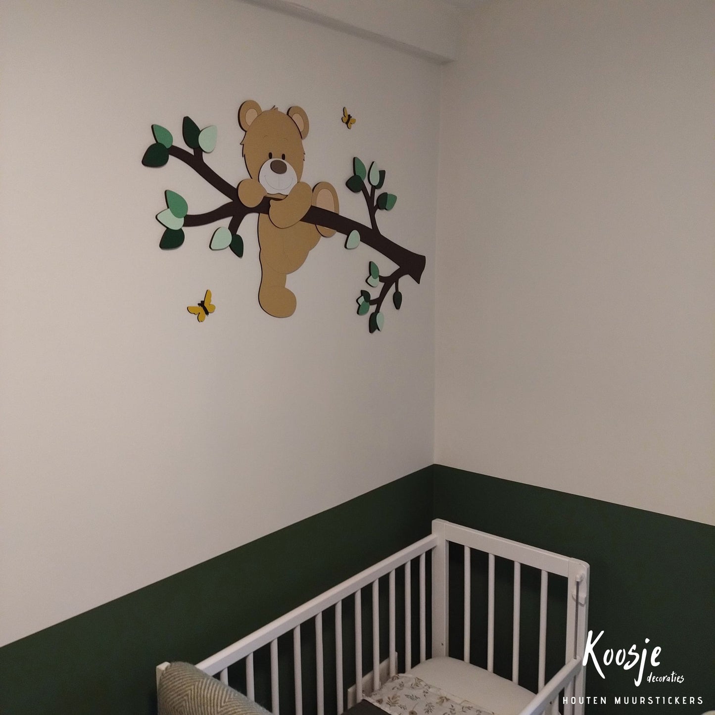 Houten decoratie babykamer met tak, groene bladeren en klauterend beige beertje boven groene lambrisering