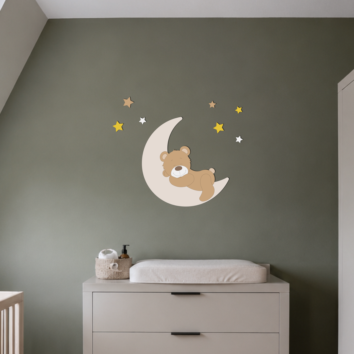 Houten muursticker voor de babykamer met een slapend beertje op een maan, omringd door sterren in geel, wit en naturel. Geplaatst boven een witte commode tegen een zachtgroene muur.

