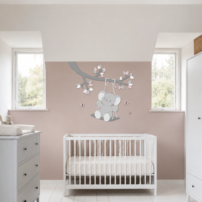 Houten muurdecoratie voor de baby- of kinderkamer met een olifantje op een schommel aan een tak, uitgevoerd in grijstinten met roze accenten, blaadjes en vlinders.