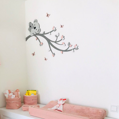 Houten muurstickers met koala op een tak boven de commode in een lichte babykamer.
De grijze tak is versierd met zachtroze blaadjes en kleine vlinders rondom de koala.
De houten muurdecoratie vormt een lieve blikvanger boven het aankleedkussen.
Inspiratie voor een roze babykamer met dieren, houten wanddecoratie en koala thema.
